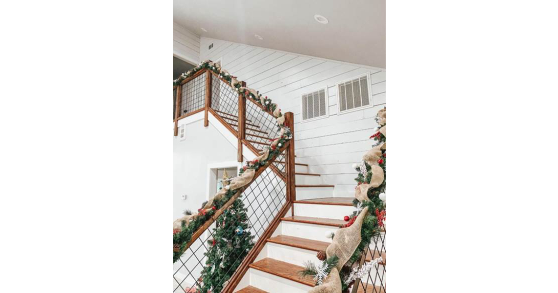 A wooden staircase in the Chapman barndominium is adorned with greenery, red ornaments, and burlap ribbon for the holidays. The white shiplap walls perfectly complement the festive decor.