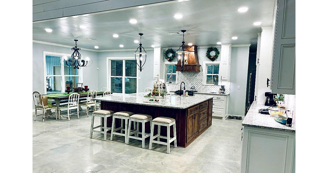 Spacious kitchen and dining area in a Chapman Barndominium with a large central island featuring a wooden base and four stools. Modern lamps and recessed lighting illuminate the space. A dining table sits near windows adorned with green wreaths, while white cabinetry lines the walls.