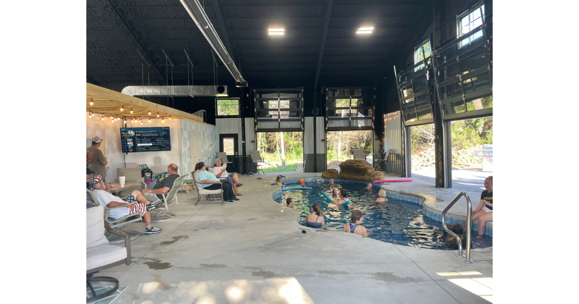The Buxton Poolhouse features an indoor pool bustling with swimmers and loungers. Large windows and a high ceiling create an airy space, where some relax on chairs and others revel in the water. In the background, a TV screen and decorative lighting enhance the serene ambiance.