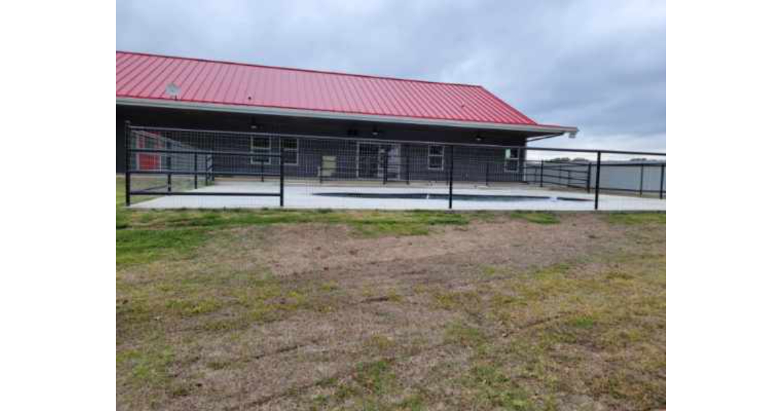 A single-story residential home with a red metal roof and dark gray exterior, surrounded by a black metal fence. The sky is overcast, and the foreground features a grassy area with patches of brown soil, reminiscent of the Freeman countryside charm.