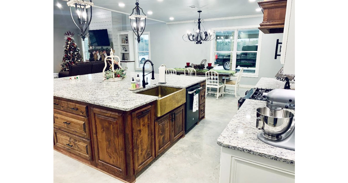 A modern kitchen in a Chapman Barndominium features a large wooden island, granite countertops, a farmhouse sink, and stainless steel fixtures. In the background, the living area showcases a Christmas tree and a dining table with white chairs, blending rustic charm with contemporary style.