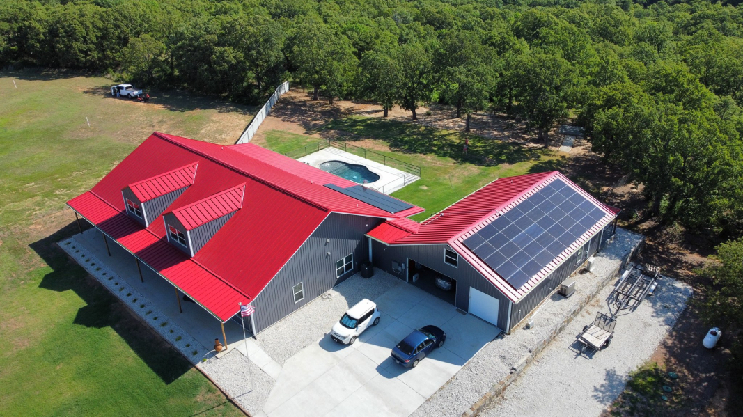 Aerial view of a modern house with red roofs and solar panels harnessing solar energy. Two cars are parked in the driveway, and a pool is visible in the backyard. The property, surrounded by greenery and woods, showcases the blend of nature and innovation.