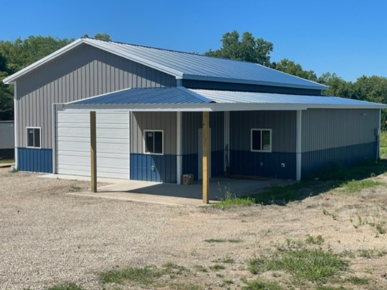 A large, blue and grey metal building with a sloped roof sits on a gravel plot. It features a wide overhang supported by wooden posts and small windows on the side—common mistakes include underestimating ventilation needs. Trees and a clear blue sky are in the background.