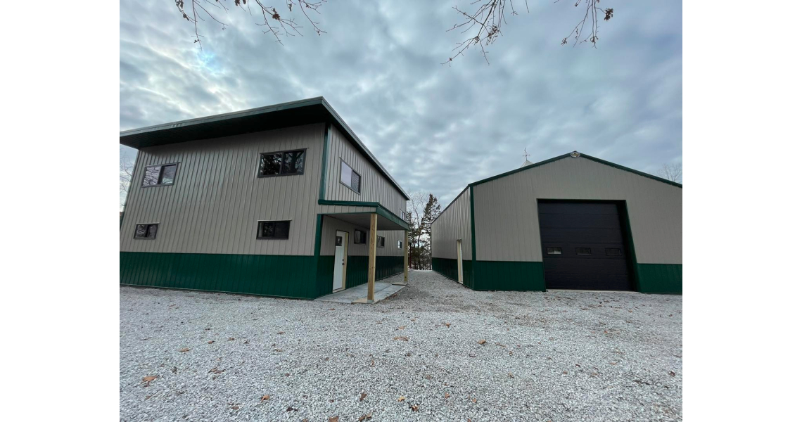 A large, modern metal barndominium with green accents and a white exterior stands on a gravel lot. Another similar structure is visible nearby under the cloudy sky, giving a glimpse of Sky Village's unique charm. Sparse trees can be seen in the background.