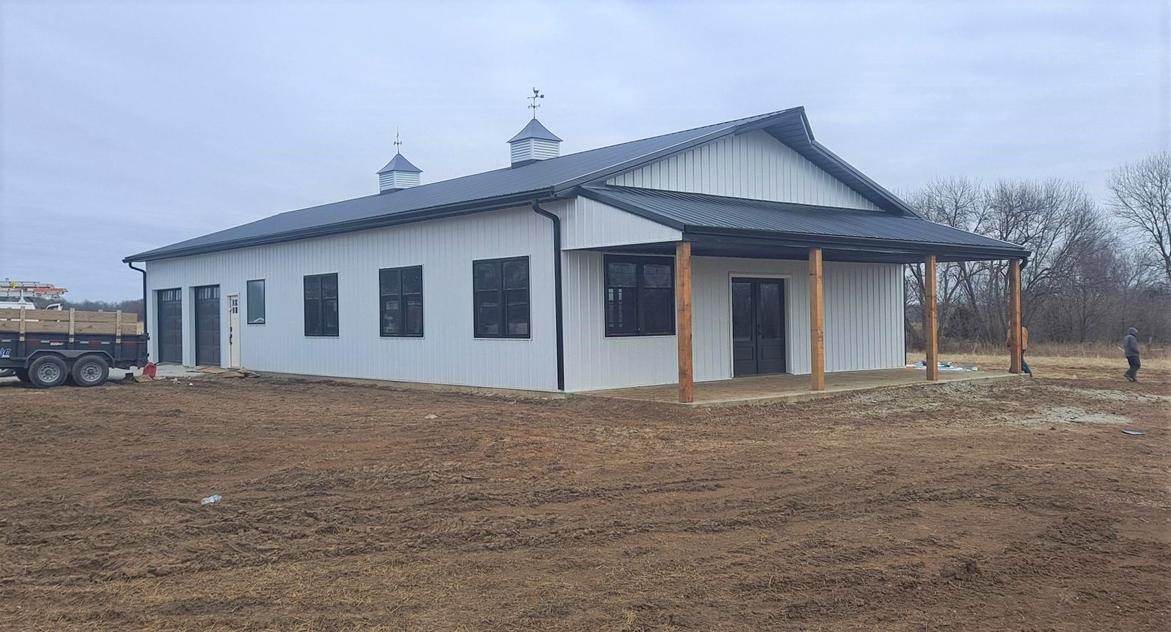 A large white barn-style building, reminiscent of a classic barndo, stands with its dark roof and wooden pillars in an open field by Pony Creek. The sky is overcast, with leafless trees silhouetted against it. Nearby, a small trailer rests while a person stands to the right.