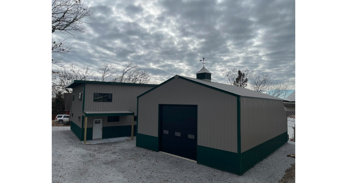 A large metal barndominium with an attached smaller structure under a cloudy sky. The building features green and gray panels, dark blue doors, and is surrounded by a gravel driveway and bare trees, evoking the charm of a rustic Sky Village.