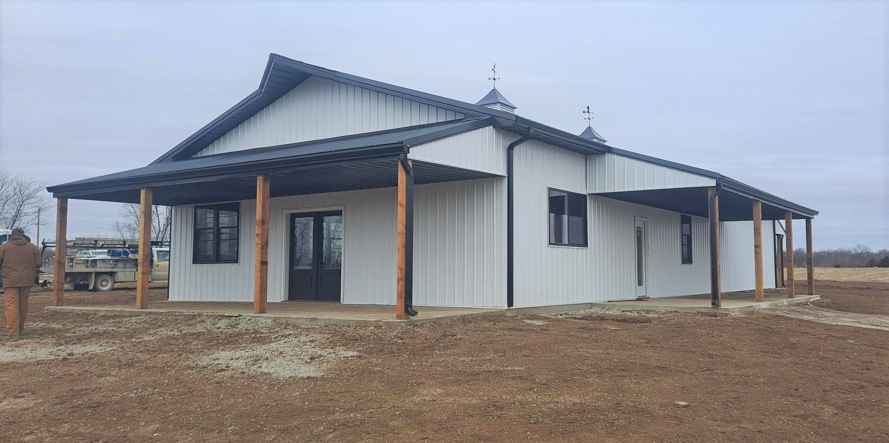 A newly constructed white metal barndo with a black roof and wooden posts supporting a wraparound porch stands proudly on a dirt lot under a gray Pony Creek sky, with a vehicle and construction materials visible in the background.