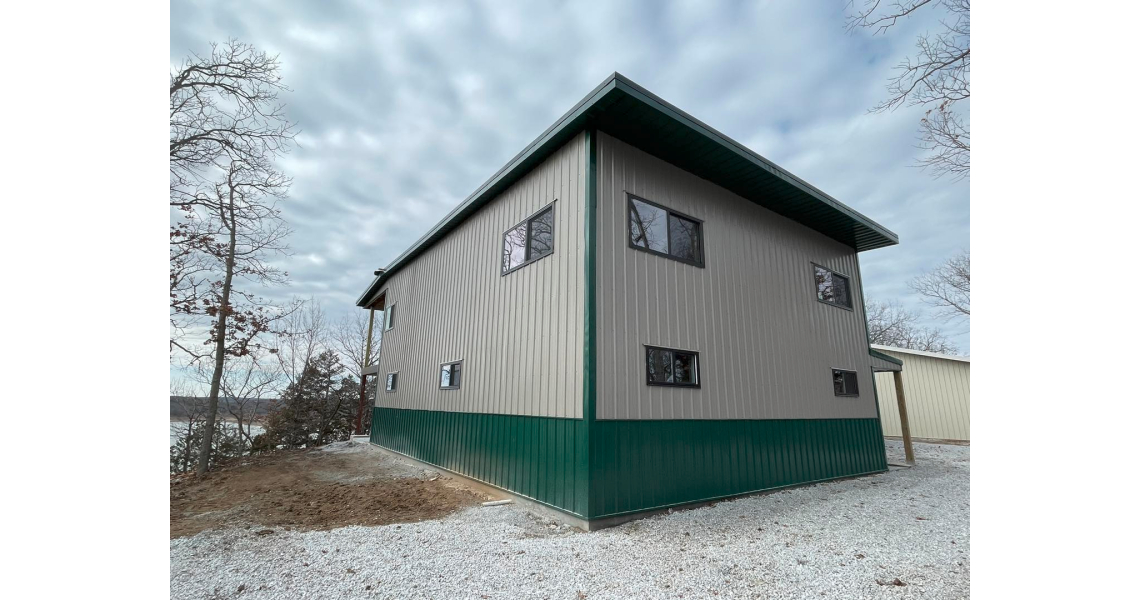 A two-story barndominium with a gray and green exterior stands on a gravel surface, embraced by leafless trees. The sky over the Sky Village is overcast with dense, gray clouds.