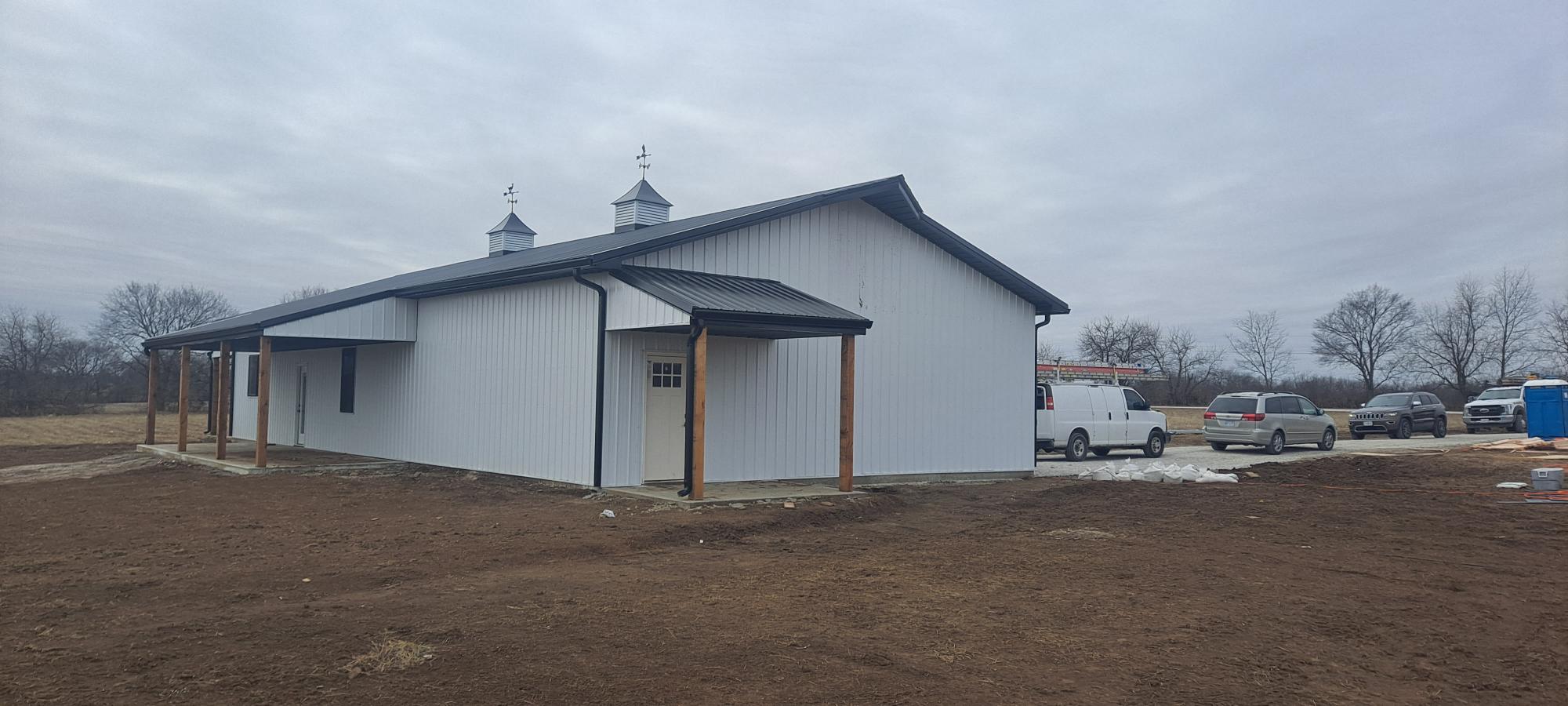 A single-story barndo with a blue roof and two small cupolas on top stands near Pony Creek. Wooden posts line its perimeter, and a few vehicles are parked nearby. The sky is overcast, casting a moody light over the largely barren landscape dotted with leafless trees.