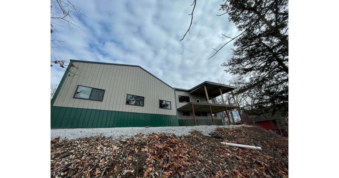 A modern, two-story barndominium with a green and beige exterior is nestled in a wooded area. The overcast sky and winter foliage envelop the Sky Village structure, which sits on a slight incline with a gravel-covered slope in the foreground.