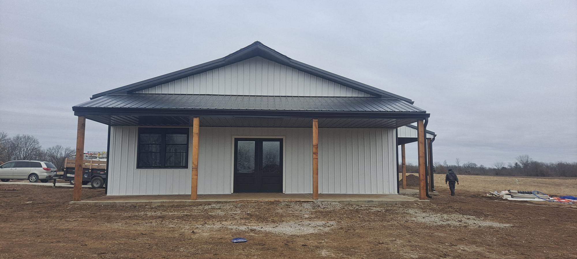 A newly constructed single-story barndo with a metal roof and wooden supports stands near Pony Creek. It features a large window and a central door. The surroundings are bare, with a car and some construction materials in the background, under an overcast sky.