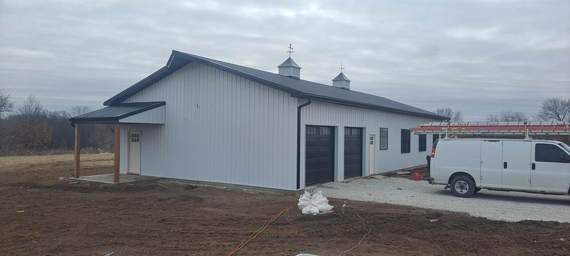 A large, modern white Barndo with a black roof and two cupolas is under construction near Pony Creek. To the right, a white van is parked next to a ladder leaning against the building. The ground is mostly dirt and gravel, with a cloudy sky above.