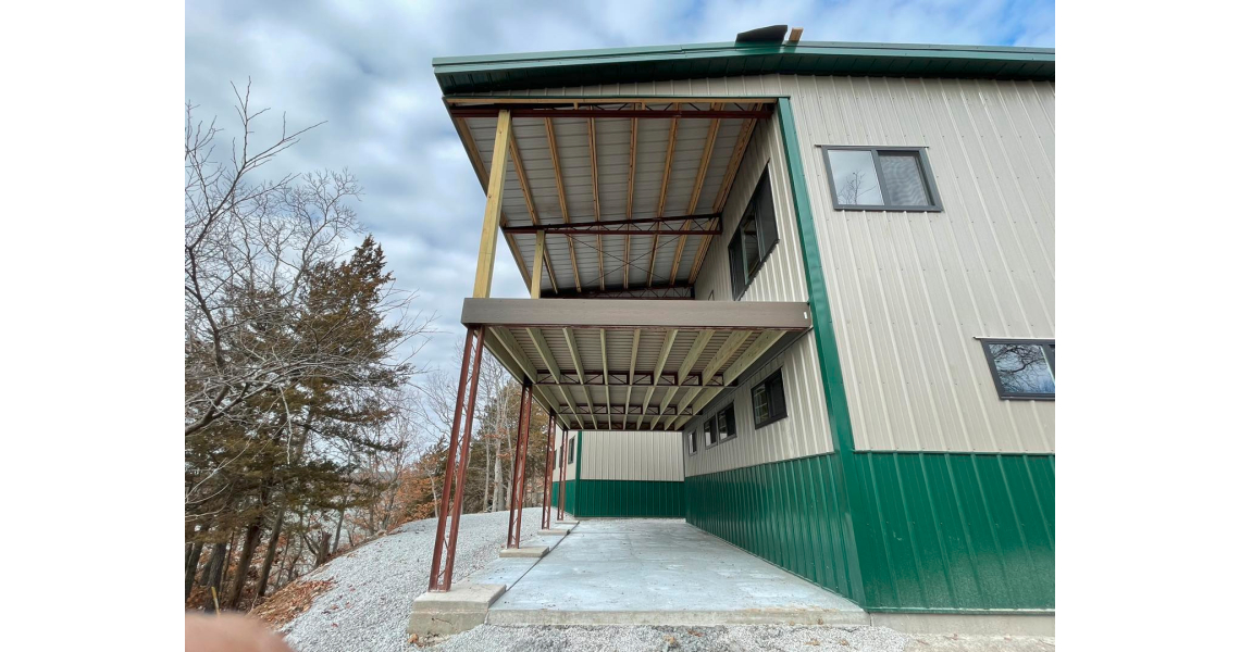 Side view of a large, two-story barndominium with a green and beige exterior. The building features a covered concrete walkway supported by beams. Surrounding the structure are trees, evoking the charm of Sky Village, with a cloudy sky in the background.