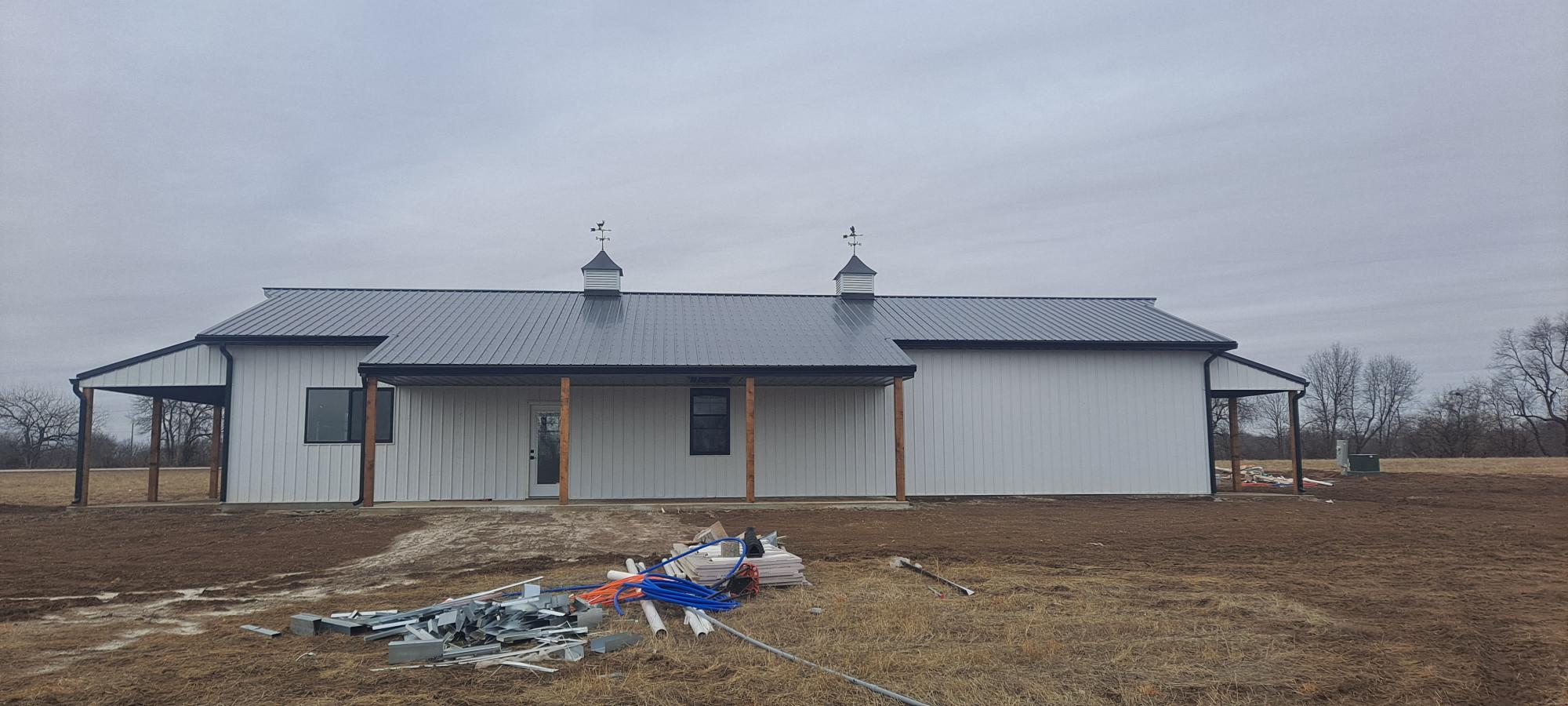 A single-story white building resembling a barndo, with a dark roof and two small cupolas, sits in a grassy field near Pony Creek under a cloudy sky. Construction materials and debris are scattered on the ground in front of the building.