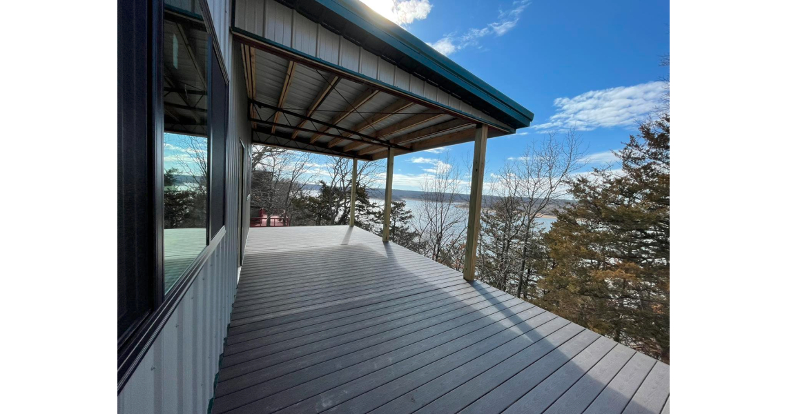 A wooden deck with a green metal roof extends from the barndominium, offering a view of a lake surrounded by trees. The sky is partly cloudy with patches of blue, creating a tranquil Sky Village vibe.