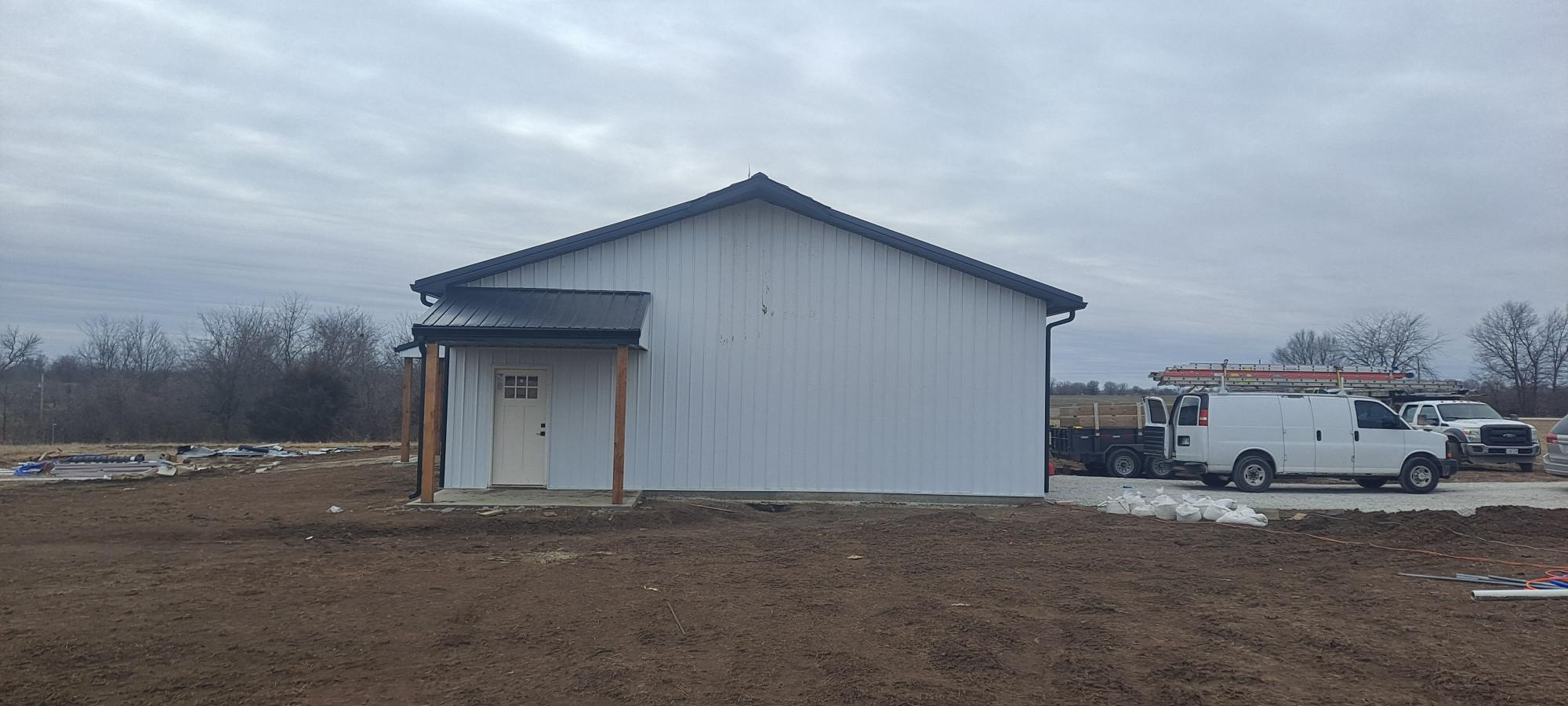 A small white building with a dark roof stands on the barren Pony Creek Barndo plot. Two wooden posts support a modest awning above a door. Nearby, two white vehicles, including a van with ladders on top, are parked under the overcast sky.