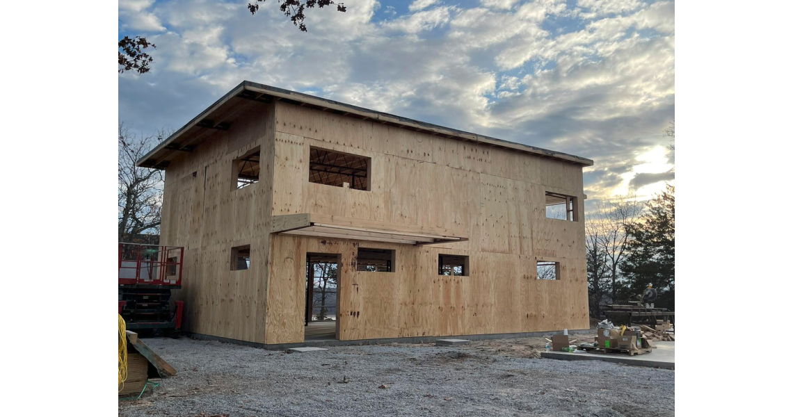A two-story wooden Barndominium stands under construction, with unfinished walls and window openings. Set on a gravel surface amidst a Sky Village of trees, the scene shows partly cloudy skies and nearby building materials.