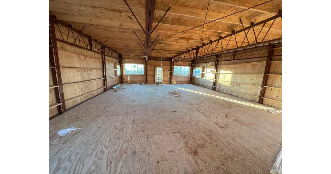 An unfinished wooden room in a rustic barndominium features a plywood floor and exposed beams. Sunlight filters through three windows, highlighting walls of plywood with visible metal supports, as construction materials lie scattered on the floor.