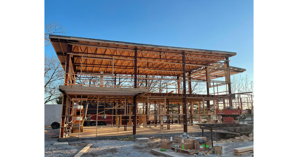 A partially constructed two-story barndominium with a visible wooden framework stands on a gravel lot. Scaffolding surrounds the structure, and construction materials are scattered around the site under a clear Sky Village sky.