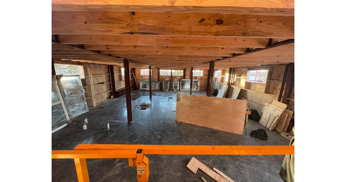 A partially constructed room in Sky Village features wooden framing on the ceiling and walls, reminiscent of a barndominium. Several windows are stacked against the walls, with loose construction materials scattered on the floor. An orange railing stands prominently in the foreground.