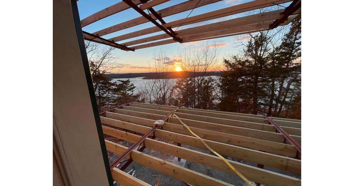 A wooden deck under construction with exposed beams overlooks a serene lake at sunset. The sky is filled with warm orange and pink hues, reminiscent of a Sky Village, while tall trees frame the view of this barndominium retreat, creating a peaceful and scenic atmosphere.