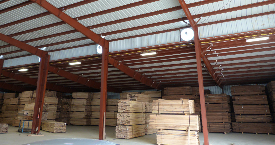 Stacks of wooden planks are neatly arranged in a spacious warehouse with a high metal roof and red support beams. This well-organized example of the different types of warehouses features ample lighting illuminating the interior.