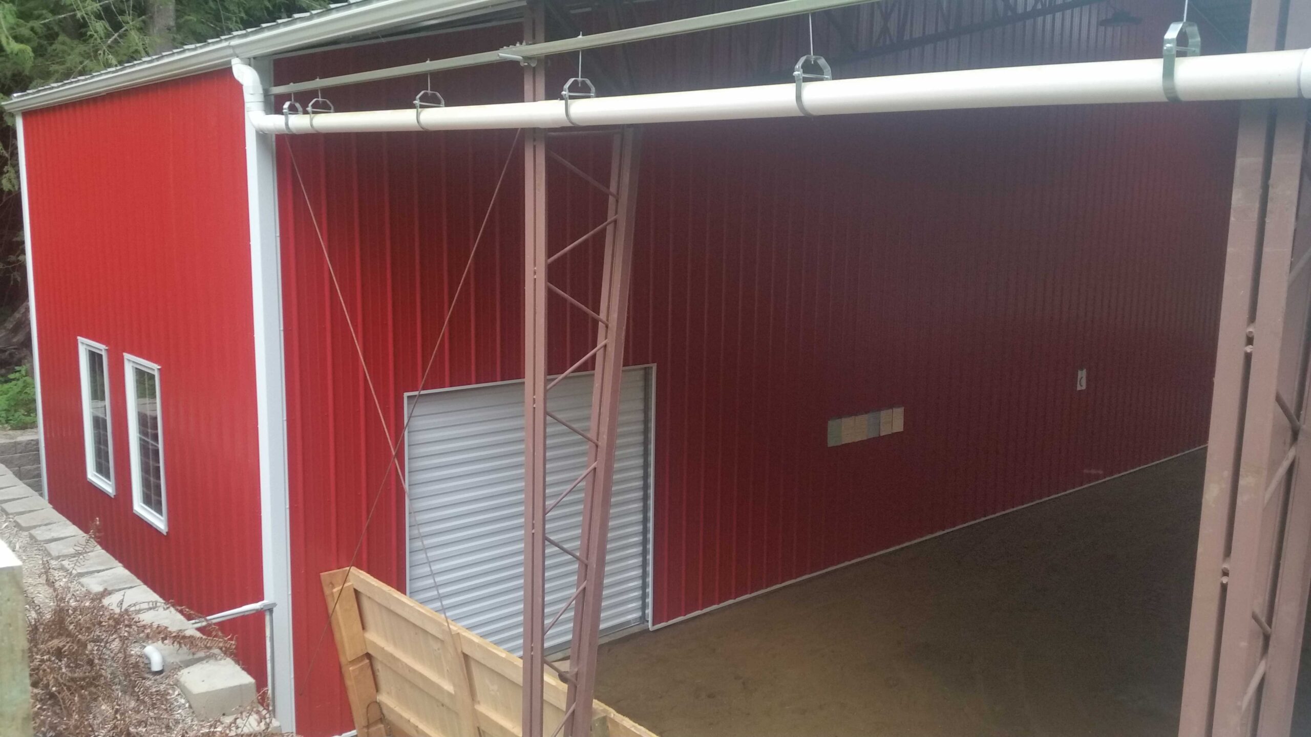 A large red metal building with a corrugated roll-up door stands prominently. Designed with a brown metal frame, the partially enclosed structure hints at its agricultural purpose. An open section reveals its interior like an arena, while a wooden gate and grassy area grace the foreground.