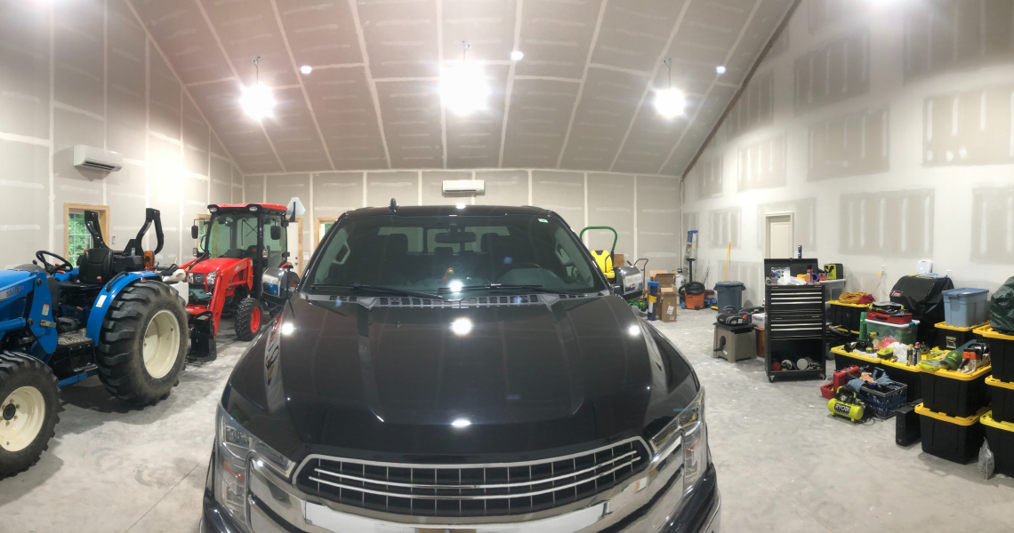 A large garage with a black truck parked in the center features shelving and toolboxes lining the walls. Red and blue tractors stand on either side, reminiscent of a Carolina Tube Leg design. Various tools and boxes are scattered throughout, while high ceilings with lights create a homey atmosphere.