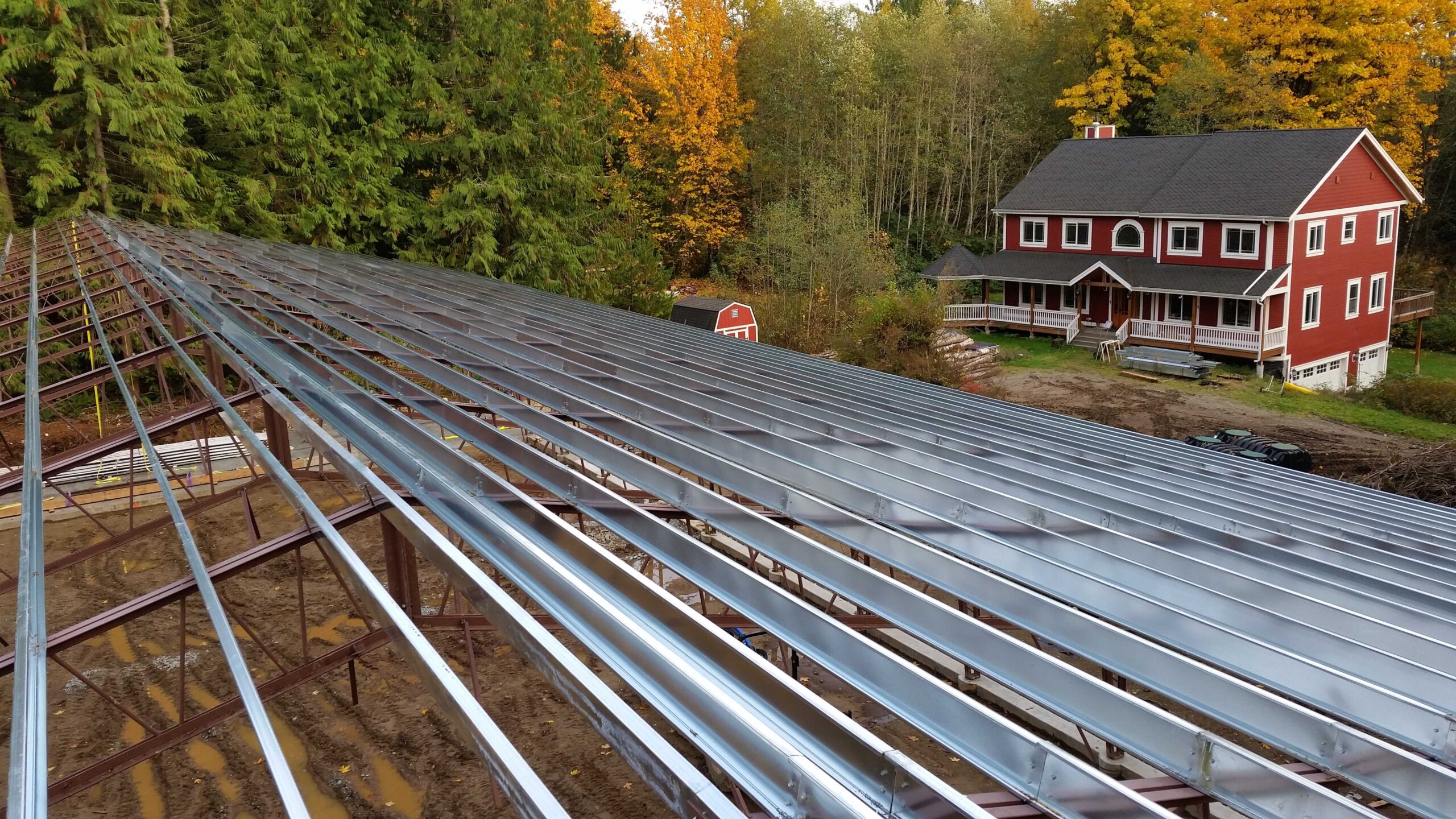A large, partially constructed metal roof, possibly for an agricultural arena, stands near a red two-story house with white trim and a smaller shed. This charming setting in the woods showcases trees adorned in vibrant autumn colors.