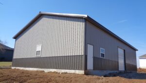 The Deer Ridge Garage is a large gray metal building with a sloped roof, resting on a dirt-covered foundation. It features a white door, a small window, and an expansive garage door. The sky is clear and blue.