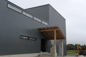 This modern metal building, rumored to be the next Volta Climbing facility, features large windows and a wooden entrance canopy supported by stone pillars. The overcast sky and greenery provide a serene backdrop, while construction equipment is visible on the right.