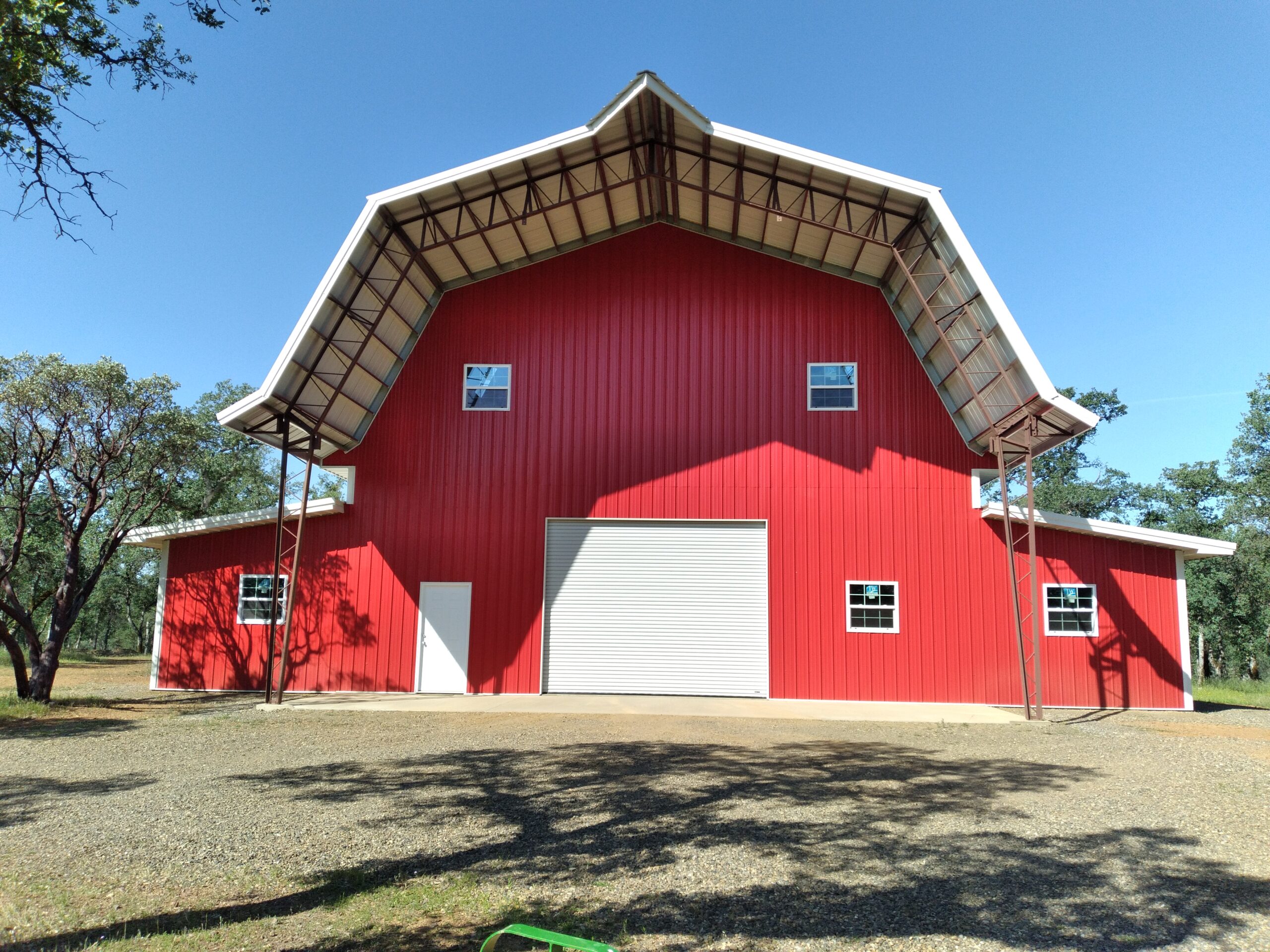 A large red barnhouse with a white roof and gambrel style stands under a clear blue sky. It features a wide central door flanked by two smaller doors and several windows. Surrounding it are trees and a gravel driveway in the foreground.