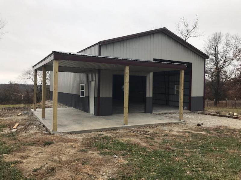 A newly constructed metal building, resembling a lodge, features an attached covered porch. Nestled on a grassy area in Hickory Hills and surrounded by bare trees, the scene exudes a gray, wintery charm under the overcast sky.