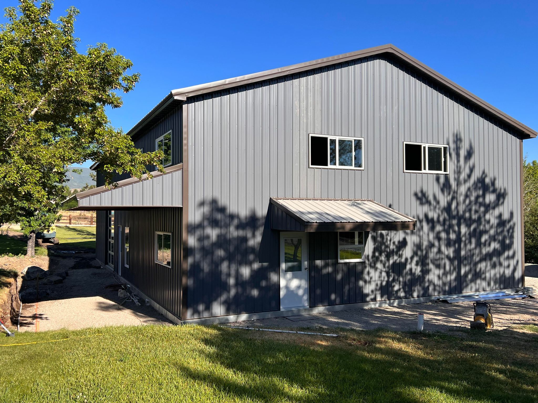 A large gray metal structure resembling a garage, with multiple windows, is nestled amid green grass and trees. It casts shadows on the ground under a clear blue sky, creating a serene river bend ambiance.
