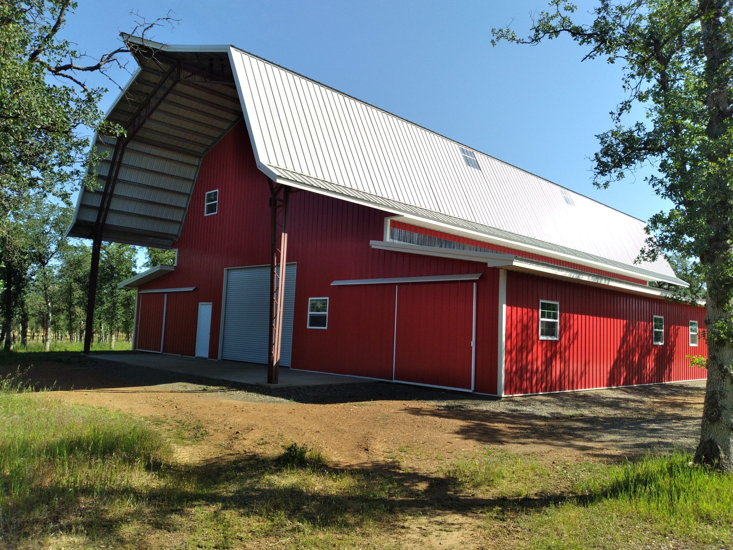 A large red barnhouse with a white metal roof, embracing the traditional gambrel style, is surrounded by grass and trees under a clear blue sky. The structure features multiple windows and a sizable sliding door on one side.