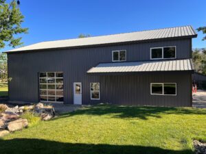 A large gray metal building with a sloped roof stands near River Bend, featuring multiple windows and a small door. A spacious garage door hints at its use as a shop. In the foreground, green grass mingles with partial landscaping of rocks and shrubs.