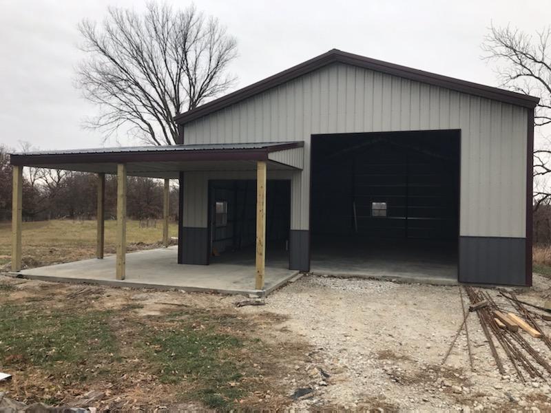 A partially constructed metal building with an open garage door and an attached covered area supported by wooden posts, Vet Lodge stands on a concrete foundation in rural Hickory Hills. Leafless trees frame this serene backdrop, hinting at future veterinary services within.