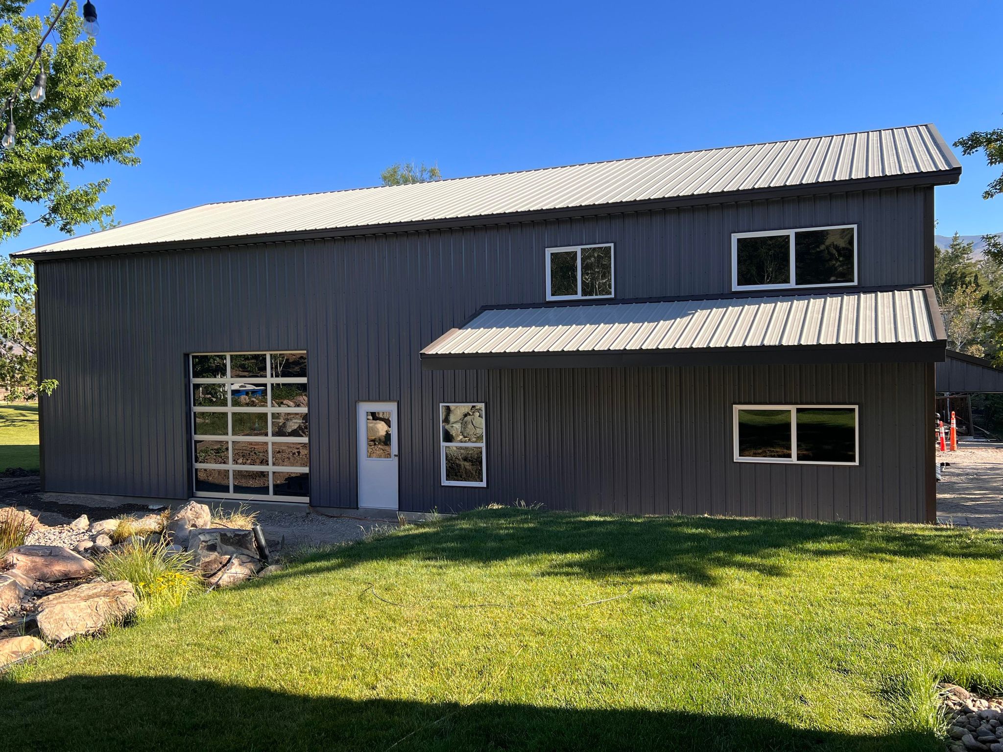 A large gray metal building with a sloped roof stands near River Bend, featuring multiple windows and a small door. A spacious garage door hints at its use as a shop. In the foreground, green grass mingles with partial landscaping of rocks and shrubs.