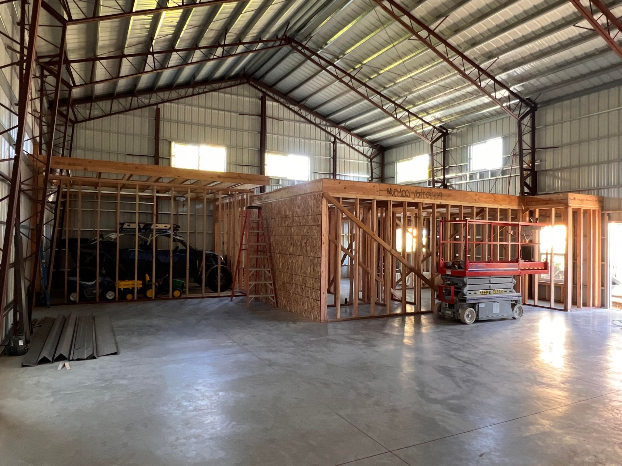 Interior of an unfinished building with a metal roof, resembling a spacious workshop. Wooden framing structures are visible, and construction equipment is present, including a scissor lift and other tools. Natural light streams in through the windows.