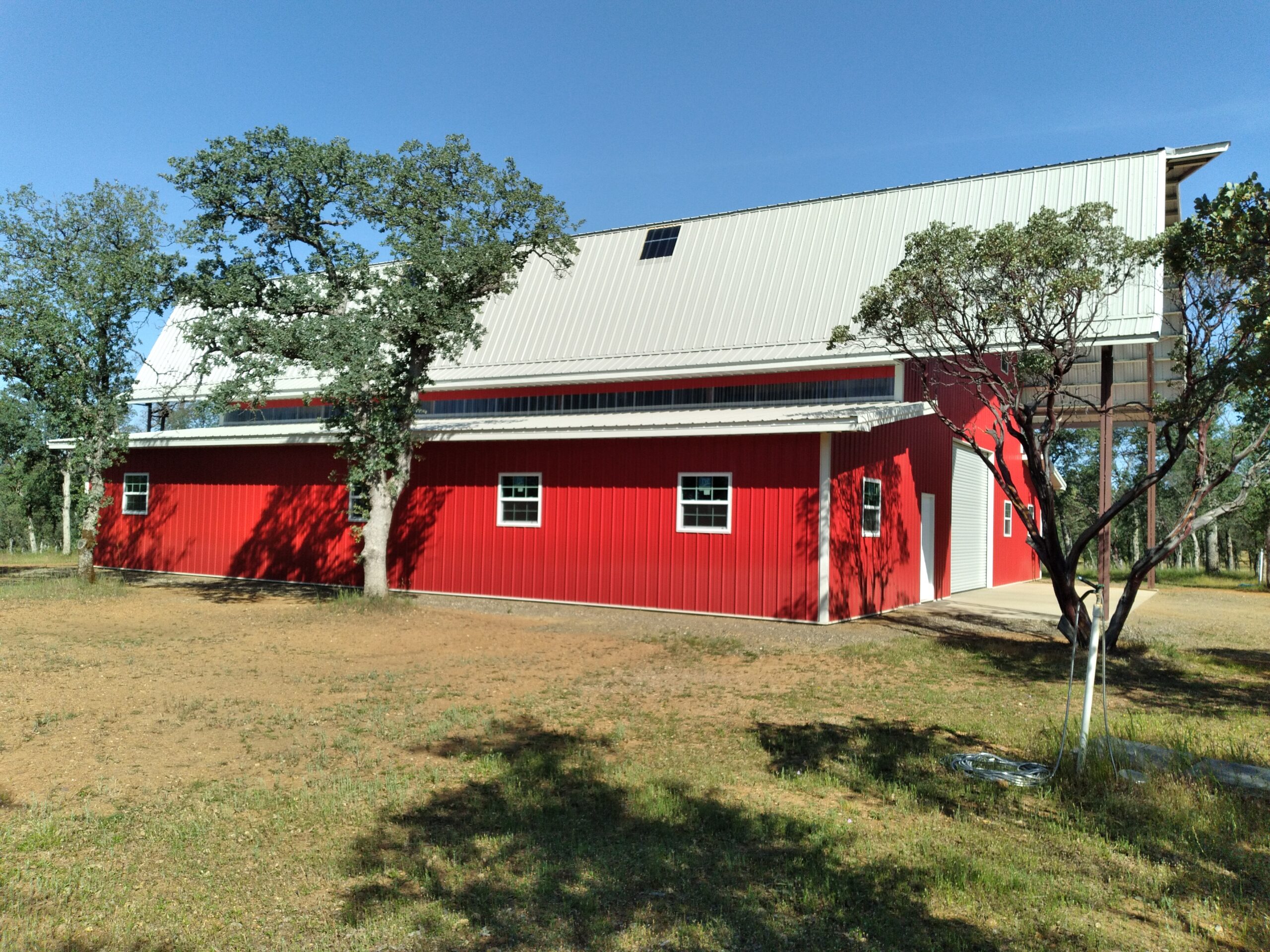 A large red barnhouse with a white gambrel roof stands in an open area under a clear blue sky. Trees and patches of grass surround the barn, with sunlight casting shadows on the ground.