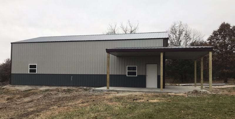 A simple, two-tone metal building with a slanted roof and the charm of a lodge. On the right side, a covered porch with wooden posts stands, reminiscent of Hickory Hills. The ground is partly grassy, dotted with bare soil patches, while trees rise in the background under a cloudy sky.