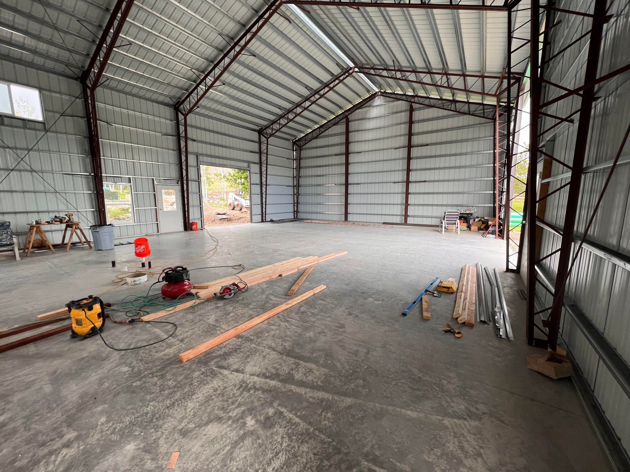 Interior view of a large, empty metal warehouse with a high ceiling, resembling an industrial garage. Various construction tools, wooden planks, and equipment are scattered across the concrete floor, with open sections revealing natural light from River Bend outside.