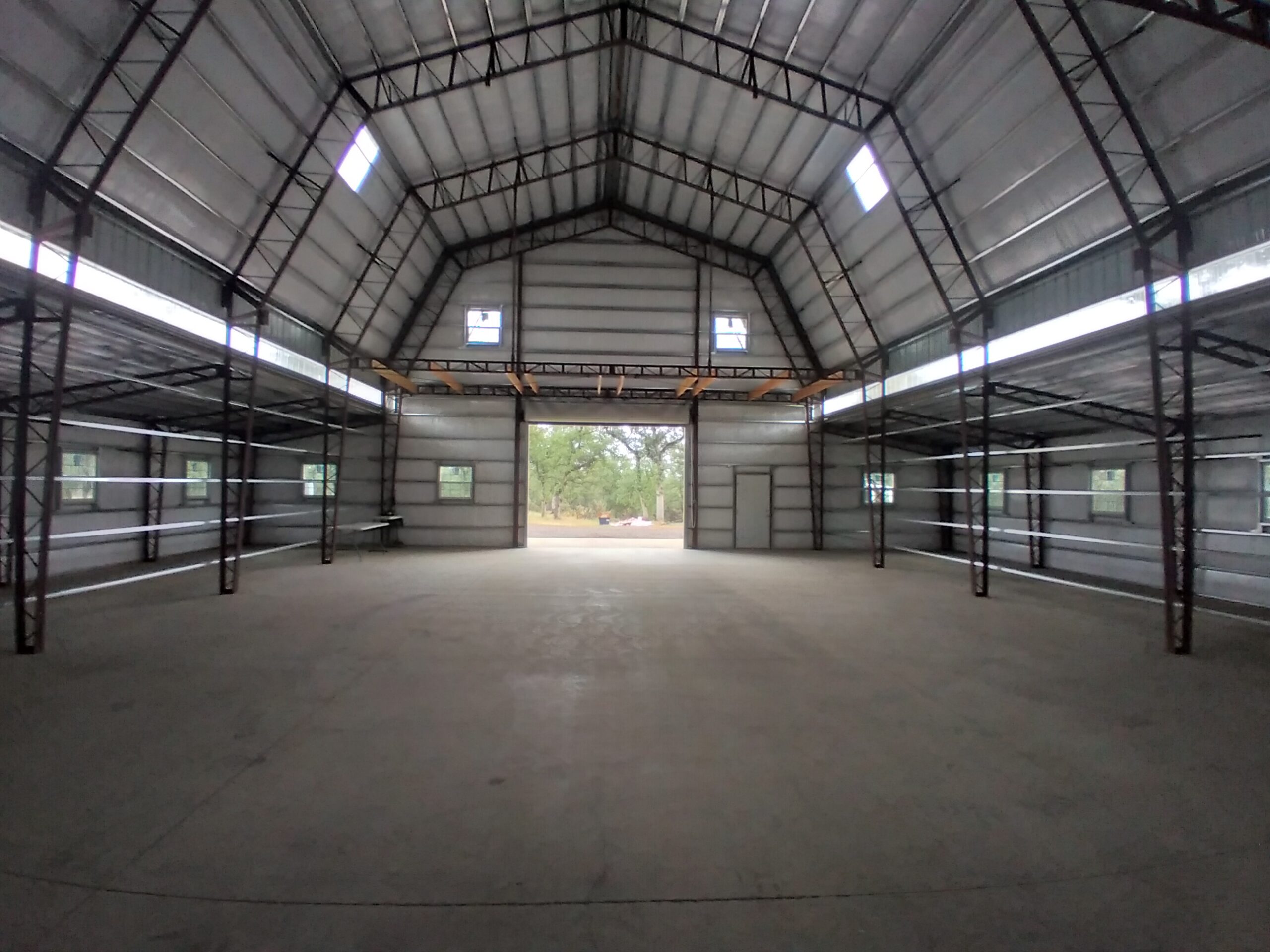 The interior of an empty warehouse features high ceilings, metal beams, and large windows. Its gambrel-style architecture adds charm to the open space, with shelving along the walls and a doorway leading outside where greenery is visible.