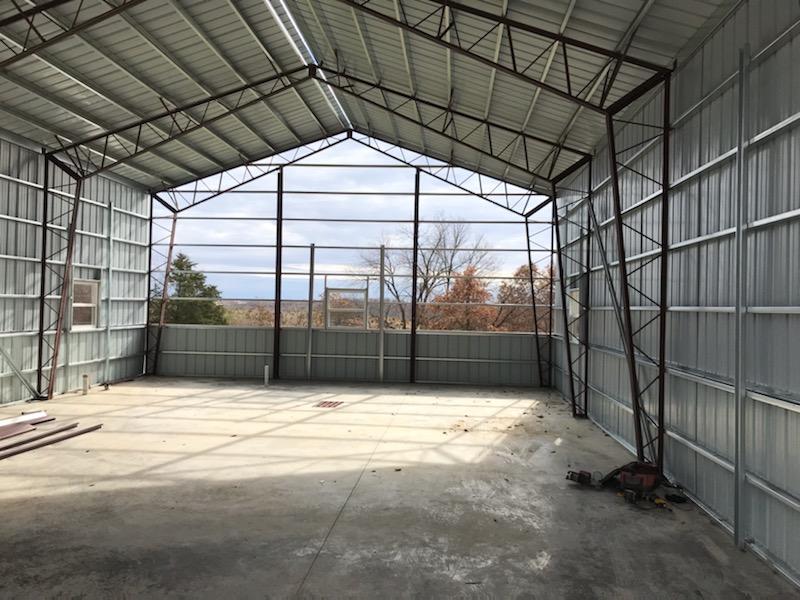 The interior of the unfinished metal-framed building at Vet Lodge boasts a concrete floor. Steel beams are visible, and large glassless windows open to a scenic view of Hickory Hills' trees and sky. Tools and materials are scattered on the ground, hinting at ongoing construction.
