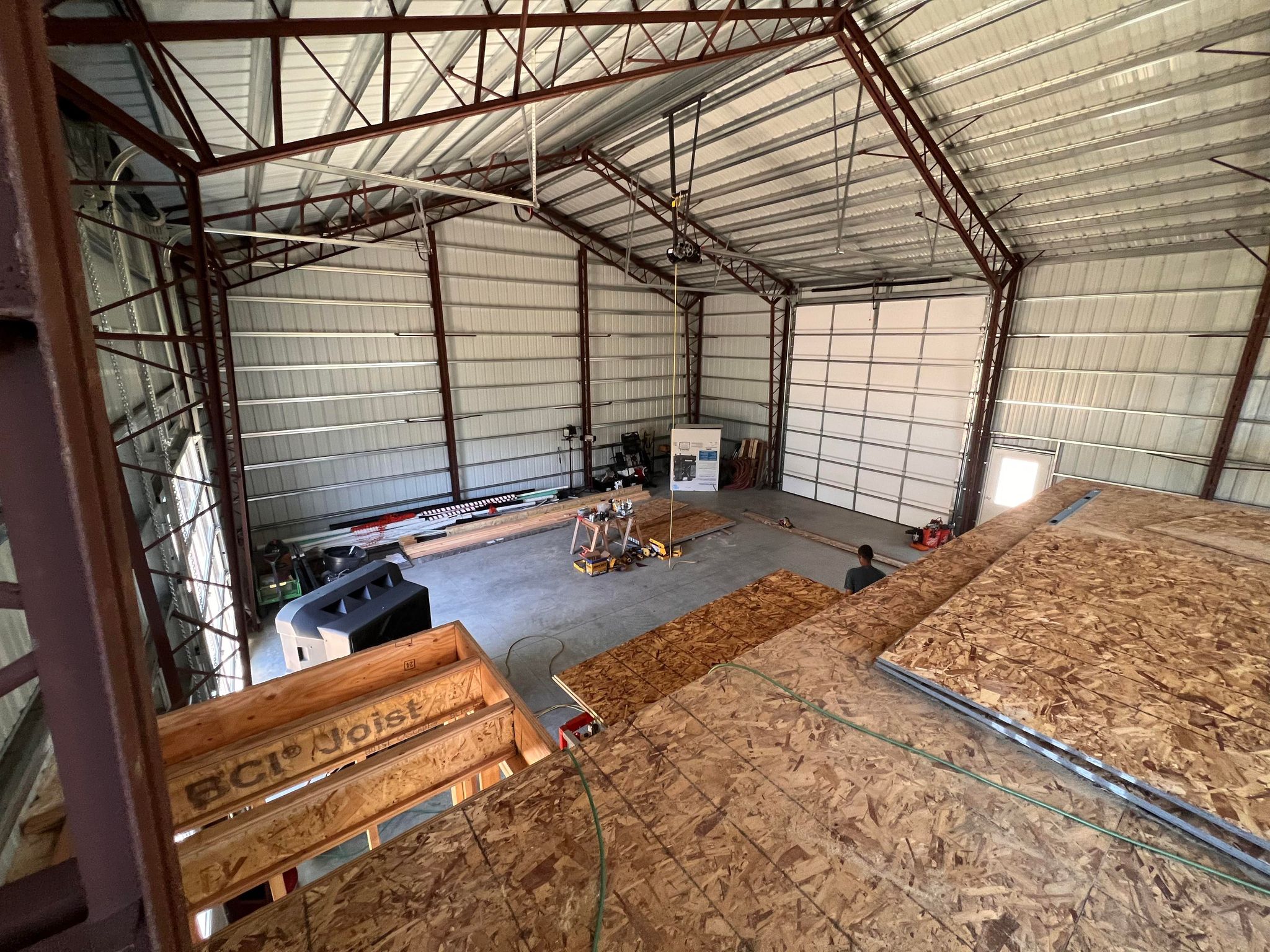 Interior view of a large metal warehouse resembles a spacious garage, with construction materials and equipment scattered around. Plywood and metal beams are visible, and a person stands near the back wall. The ceiling is high with structural supports.