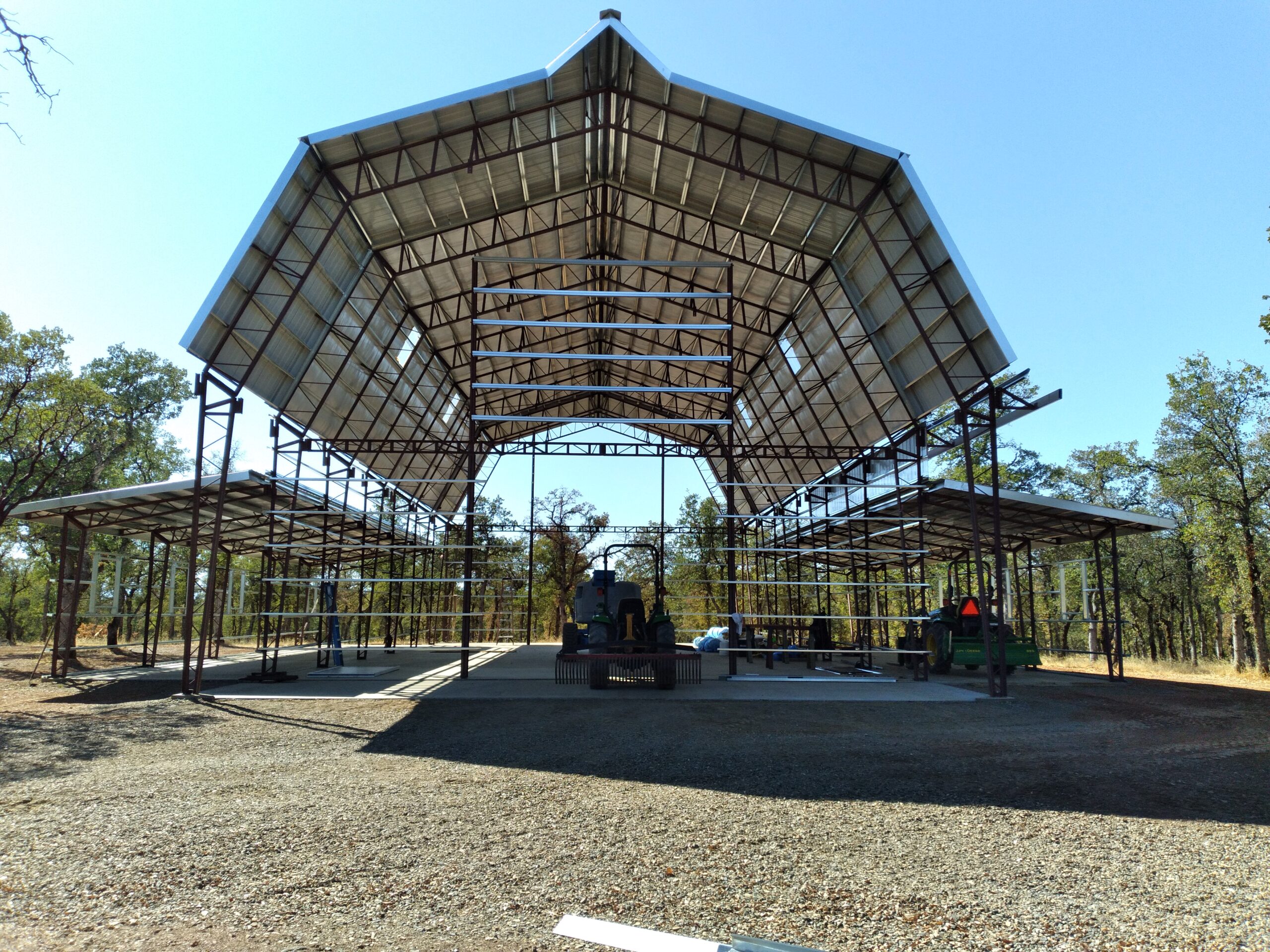 A large open metal structure with a pointed gambrel-style roof, partially built on a gravel lot. Construction equipment is visible inside the barnhouse, surrounded by trees under a clear blue sky.