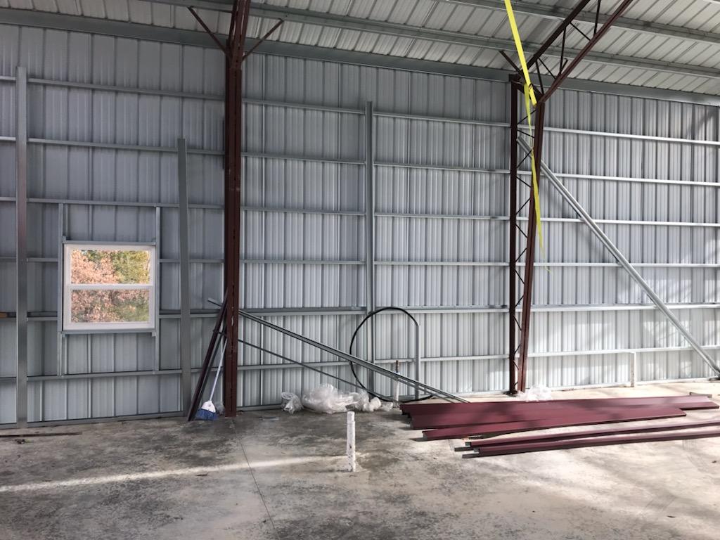 Interior view of a lodge under construction, featuring corrugated steel walls and beams. Several steel panels rest on the concrete floor alongside construction materials. A window on the left reveals trees in the scenic Hickory Hills outside.