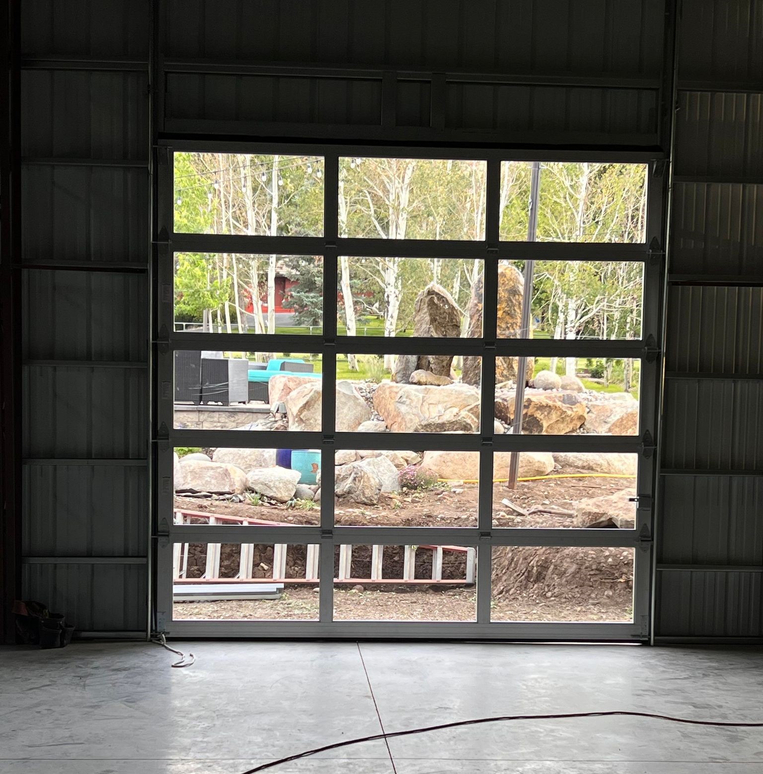 A large, open garage door reveals a view of a yard with rocks and trees. In the background, several large boulders sit amidst deciduous trees with green leaves. Inside this spacious shop-like garage, you'll find a concrete floor and metal walls.