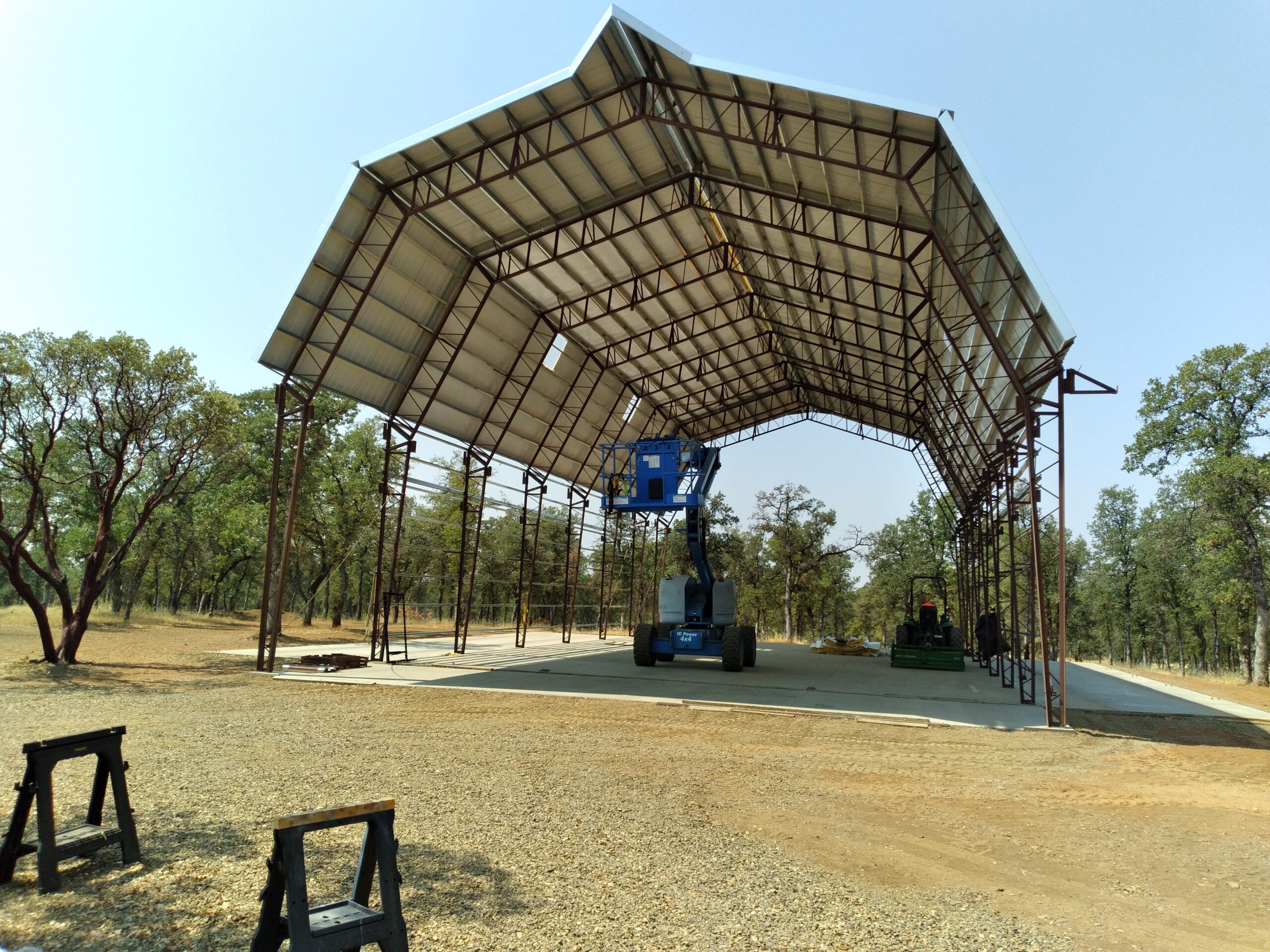 A large gambrel-style metal structure is under construction, resembling a hangar or storage shed. A blue cherry picker is lifting a person inside. Trees are visible in the background on a sunny day, surrounded by dry, gravelly ground.