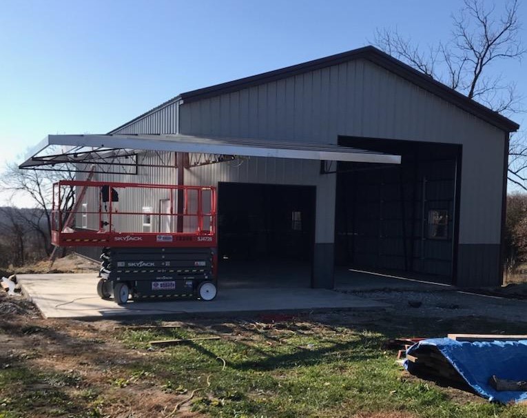 A metal building with large garage doors is under construction in Hickory Hills. An elevated lift with a worker is stationed at the entrance, installing an awning. The ground is covered with dirt and construction materials, while trees and a clear sky form the backdrop.